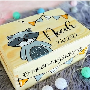 May include: A wooden keepsake box with a raccoon illustration, the name "Noah" and the date "24.12.2022" written on it. The box is decorated with a string of colorful flags and the text "Erinnerungskiste" which translates to "memory box".