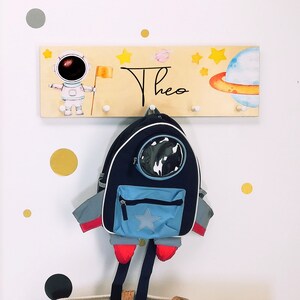 Könnte beinhalten: Ein Holz-Kleiderständer mit Weltraum-Motiv. Er zeigt einen Astronauten, Planeten, Sterne und den Namen "Theo". Ein blauer und schwarzer Rucksack mit Raketen-Design hängt am Kleiderständer.