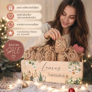 Peut inclure: Une caisse en bois marquée Lauras Adventskalender avec une illustration de village festif et des ornements verts. Elle contient plusieurs petits sacs en toile de jute à cordon, chacun orné d'une étiquette numérotée en bois.