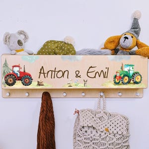 Peut inclure: Porte-manteau mural en bois avec crochets, présentant un design personnalisé avec les noms Anton & Emil, deux tracteurs jouets et un paysage vert, bleu et blanc.