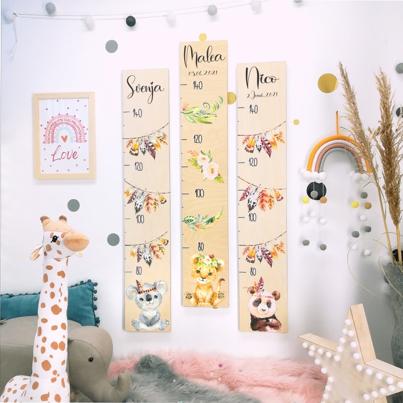 May include: Three wooden growth charts with a boho theme. Each chart features a different animal: a koala, a lion, and a panda. The charts are decorated with feathers, leaves, and flowers. The charts are labeled with the names "Svenja", "Malea", and "Nico" and their birth dates.