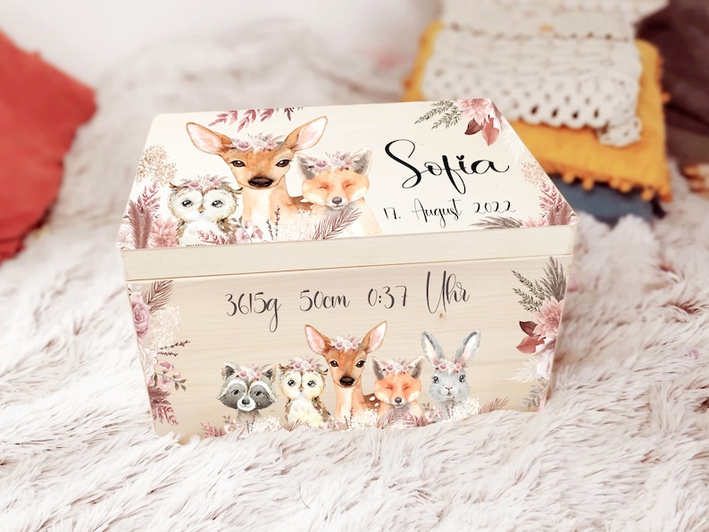 K&ouml;nnte beinhalten: Eine h&ouml;lzerne Erinnerungsbox mit hellbraunem Hintergrund und floralen Akzenten. Die Box zeigt eine fantasievolle Waldlandschaft mit einem Reh, einer Eule, einem Fuchs und einem Waschb&auml;r. Die Box ist personalisiert mit dem Namen "Sofia", dem Datum "17. August 2022" und den Geburtsdaten "3615g 50cm 0:37 Uhr".