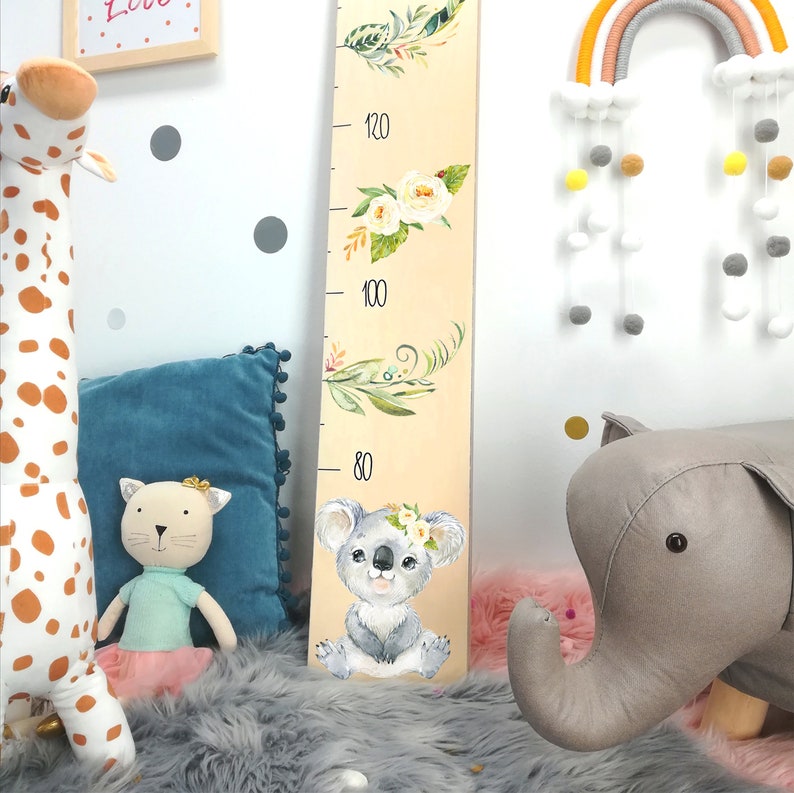 May include: A wooden growth chart with a light brown background and a white koala bear with a floral crown. The chart has numbers from 80 to 120 printed on it. The chart is decorated with watercolor floral designs.