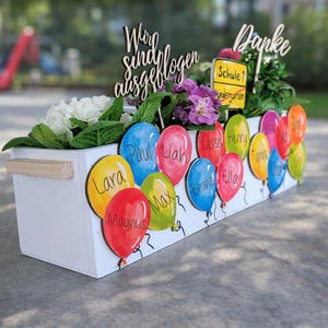 Könnte beinhalten: Ein weißer Holzblumenkasten, verziert mit bunten Ballonausschnitten, auf denen jeweils der Name eines Kindes steht. Die Box ist mit leuchtenden Blumen und Grün gefüllt, mit Holzschildern mit der Aufschrift "Danke" und "Wir sind ausgeflogen."