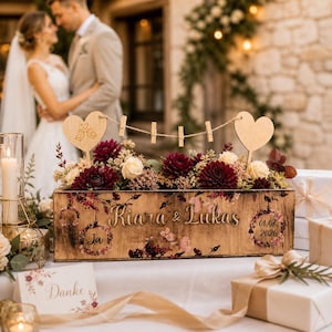 Op de afbeelding: Een houten bloemstuk met de namen "Kiara & Lukas" en de datum "01.08.2026" gegraveerd. Het heeft bordeauxrode dahlia's, crèmerozen en een hartvormig houten accent. Een bruid en bruidegom zijn wazig op de achtergrond.