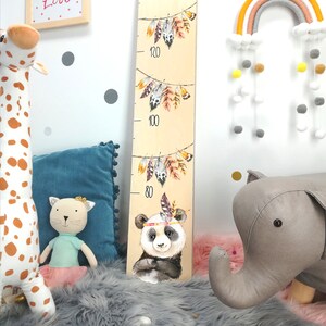 May include: A wooden growth chart with a panda bear, feathers, and the numbers 80, 100, and 120. The chart is on a white wall with a gray and white pom-pom garland.