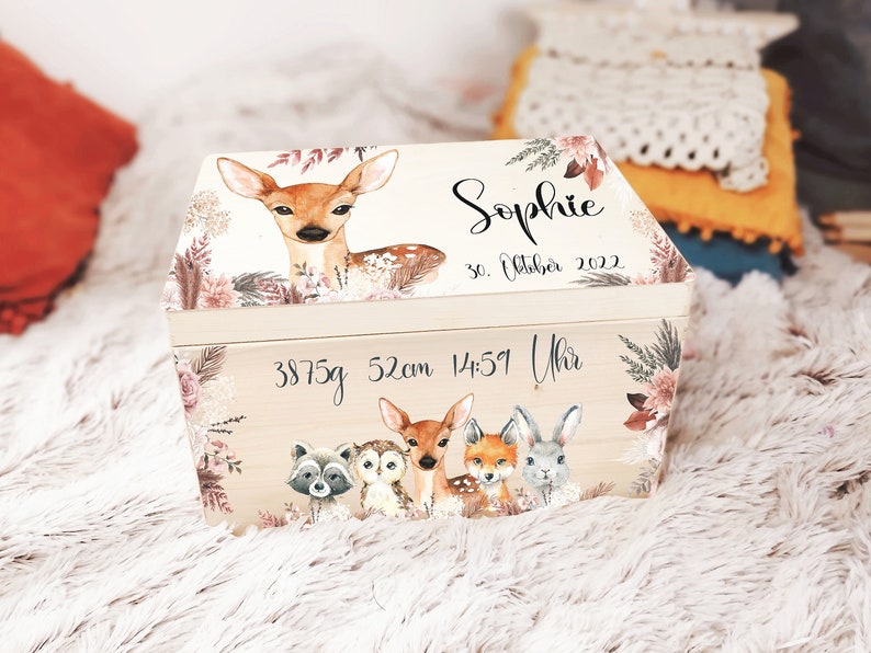 K&ouml;nnte beinhalten: Eine Holz-Erinnerungskiste mit einer hellen Holzoptik und einem floralen Design. Die Kiste hat einen Klappdeckel und ist mit einem Reh, einer Eule, einem Waschb&auml;r, einem Fuchs und einem Hasen verziert. Die Kiste ist personalisiert mit dem Namen "Sophie", dem Datum "30. Oktober 2022" und der Uhrzeit "14:51 Uhr". Die Kiste enth&auml;lt auch die Ma&szlig;e "3875g 52cm".