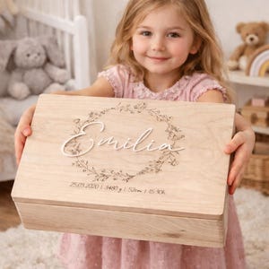 Op de afbeelding: Een lichtgekleurde houten herinneringskist met de naam "Emilia" in script, omringd door een bloemenkrans. De doos bevat ook de datum 25.03.2020, gewicht 3480 g, lengte 52cm en tijd 15:30h.
