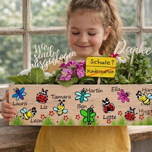 Op de afbeelding: Een houten plantenbak met geschilderde insecten en bloemen, vastgehouden door een persoon. De doos heeft namen en de woorden "Schule" en "Kindergarten" op een bord. De doos is gevuld met planten en bloemen.