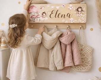 May include: A light wood wall-mounted coat rack featuring a shelf with a printed design of mushrooms, fairies, and the name Elena. The rack includes four wooden pegs holding a beige hooded jacket, a pink quilted jacket, and a crochet bag.