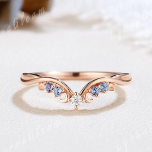May include: Rose gold ring with a curved design. The ring features a central white gemstone and several blue gemstones. The ring has a delicate, elegant appearance, suitable for special occasions.