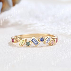 May include: A gold ring with a delicate band, set with a variety of colourful gemstones. The ring features a cluster of stones in different shapes, including round, oval, and rectangular. The gemstones are in shades of blue, pink, yellow, and green, creating a vibrant design.