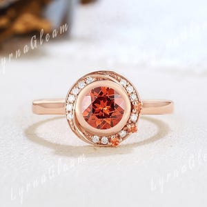 May include: Rose gold ring with a central, round, fiery orange gemstone, surrounded by small white diamonds. The ring also features additional orange gemstones. The band is delicate, and the setting is halo-style.