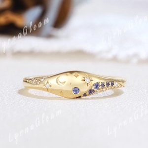 May include: A gold ring with a celestial design. The ring features a crescent moon, stars, and small blue and white gemstones. The ring has a unique, curved shape and is set on a white surface.
