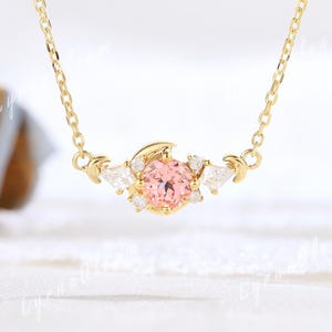 May include: A delicate gold necklace featuring a central pink gemstone surrounded by smaller white stones. The necklace has a crescent moon design on each side of the pink stone. The chain is a fine gold link.