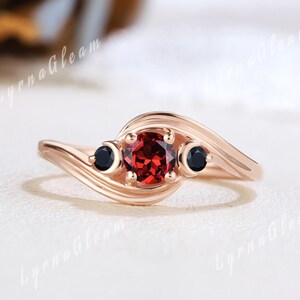 May include: A rose gold ring featuring a central round, faceted red gemstone flanked by two smaller black gemstones. The band has a swirling design, and the ring is set against a soft, neutral background.
