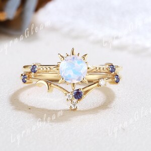 May include: A gold-colored ring set featuring a celestial design. The engagement ring has a large, round, opalescent stone surrounded by a sunburst design. The band is adorned with small, dark blue stones. The accompanying band has a curved design with additional blue stones and small white stones.