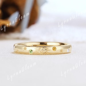May include: A gold ring with celestial designs, including a sun, moon, and stars. The ring features small gemstones in green, yellow, and clear colours. The ring is on a white surface.