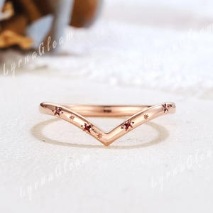 Puede incluir: Un anillo en forma de V de oro rosa con pequeñas piedras preciosas rojas y grabados en forma de estrella. El anillo tiene un diseño delicado, con las piedras preciosas engastadas en las puntas de la V. Es una joya delicada.