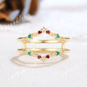 May include: A gold-colored ring guard with a decorative design. The ring features a series of small, round, red and green gemstones. The ring guard is designed to be worn with another ring. The ring guard is a jewelry item.