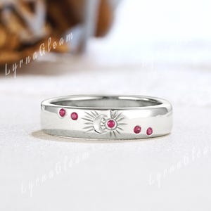 May include: A silver ring with a celestial design. The ring features a sun, moon, and starburst pattern, with small red gemstones. The ring is a band style, with a polished finish.