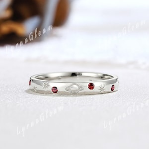 May include: A silver ring with a celestial design. The ring features a series of small, round, red gemstones interspersed with engraved stars and a planet. The ring is set against a white background.
