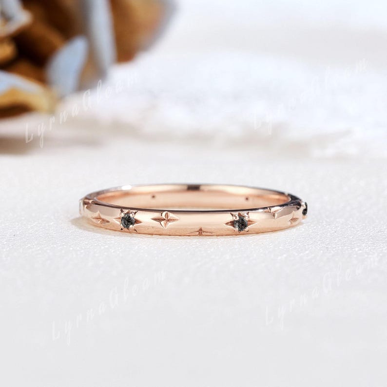 Puede incluir: Un delicado anillo de banda de oro rosa con un sutil patr&oacute;n de estrellas y peque&ntilde;os diamantes negros engastados en el borde.