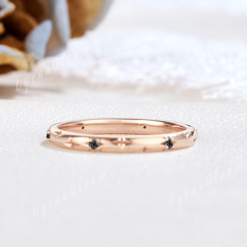 Puede incluir: Un delicado anillo de banda de oro rosa con un dise&ntilde;o texturizado y diamantes negros. El anillo est&aacute; sobre un fondo blanco.