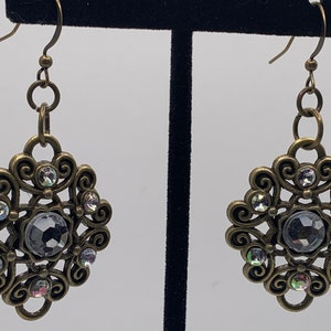 May include: A pair of gold-toned earrings with a large, clear crystal in the center of each earring. The crystal is surrounded by smaller, clear crystals and the earrings have a delicate, ornate design.