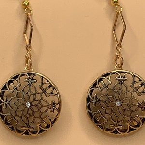 Può includere: Un paio di orecchini dorati con un design delicato e intricato. Ogni orecchino presenta un pendente rotondo traforato con un piccolo strass trasparente al centro.