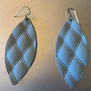 May include: A pair of pale blue, leaf-shaped earrings featuring a quilted design. The earrings have a silver-coloured hook closure. The quilted pattern is formed by raised, dotted lines, creating a diamond pattern across the surface.