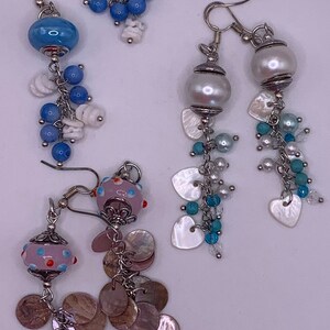 May include: Three pairs of dangle earrings with different coloured glass beads and charms. The first pair has blue glass beads and white shell charms. The second pair has white pearl beads and white shell heart charms. The third pair has pink and white glass beads and brown shell coin charms.
