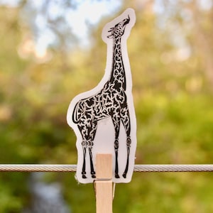 May include: A black and white illustration of a giraffe printed on a white sticker. The sticker is attached to a wooden clothespin and hanging from a wire.