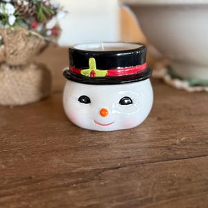 May include: A ceramic snowman candle holder with a black top hat, red ribbon, and a smiling face. The candle holder has a white body with black eyes, a carrot nose, and a red smiling mouth. A small candle sits inside the holder.