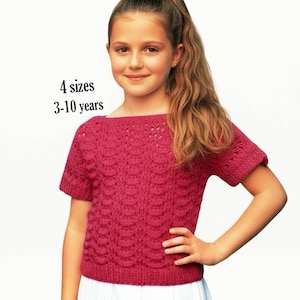 May include: A short-sleeved, magenta-colored knit top featuring a textured, repeating shell-like pattern across the front. The garment has a boat neckline and ribbed edges. Text on the image indicates 4 sizes available for ages 3-10 years.