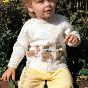 Puede incluir: Un suéter de punto blanco con un patrón multicolor y un diseño de conejo marrón en la parte delantera. El suéter lo lleva un niño sentado en un muro de ladrillo.
