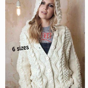 Op de afbeelding: Een gebroken wit, grof gebreid vest met capuchon, voorzien van een kabelpatroon, een knoopsluiting aan de voorzijde met vijf ronde knopen en lange mouwen. De tekst 6 sizes staat in het zwart gedrukt op de linkerzijde van het gebreide kledingstuk.