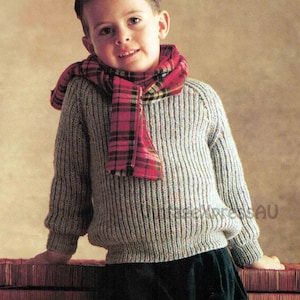 May include: A young boy wearing a grey knitted jumper with a red and black tartan scarf. He is standing in front of a wooden chest.
