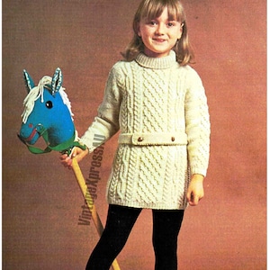 May include: A young girl wearing a white cable knit sweater dress with a brown belt and black tights. She is holding a blue and white toy rocking horse.