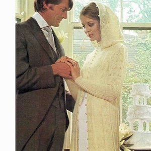 May include: A couple holding hands during a wedding ceremony. The bride is wearing a white knitted hooded dress with pearl embellishments. The groom is wearing a dark suit and a white tie.