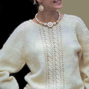 May include: A white knitted jumper with a delicate cable knit pattern and a round neckline. The jumper features a wooden bead necklace with a large, round pendant.