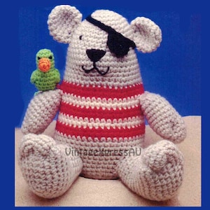 May include: Handmade crocheted pirate bear toy, featuring a black eye patch, red and white striped top, and a small green parrot. The gray bear sits on a sandy surface. A charming, handcrafted item.