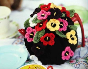 May include: A black teapot adorned with colorful crocheted flowers in shades of red, pink, yellow, and black, with green leaves. The teapot has a red handle and spout, and is set on a floral patterned tablecloth.