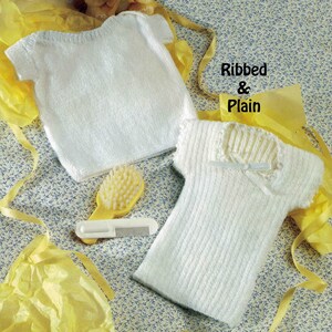 May include: Two white knitted baby shirts, one plain and one ribbed, with a small yellow comb and brush set. The text "Ribbed & Plain" is visible in the image. The items are arranged on a floral patterned surface with yellow tissue paper.