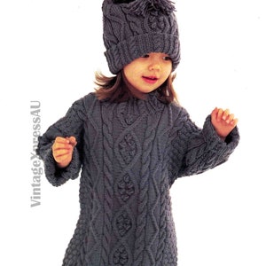 Peut inclure: Une robe pull en tricot gris avec un motif en tricot torsadé. La robe a des manches longues et un col rond. Le mannequin porte un bonnet en tricot gris avec un pompon sur le dessus.