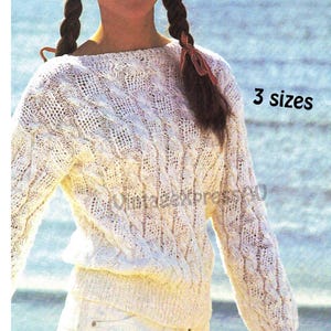 May include: A white, long-sleeved sweater with a textured knit pattern. The sweater has a crew neck and a fitted waist. The image includes the text "3 sizes". The background is a blurred view of the ocean.