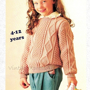 May include: A pink knitted sweater with a cable knit pattern and a textured stitch. The sweater is for ages 4-12 years.