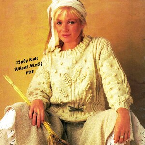 Op de afbeelding: Een witte gebreide trui met een tarwe motief. De trui heeft een ronde hals en lange mouwen. De trui wordt gedragen door een persoon met blond haar en een wit hoofdband. De tekst "12ply Knit Wheat Motif PDF" is zichtbaar in de afbeelding.