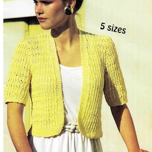 Könnte beinhalten: Ein kurzärmeliger, offener gelber Cardigan. Das Strickkleid wird über einem weißen Kleid getragen. Der Text "5 Größen" ist sichtbar. Der Cardigan hat ein strukturiertes, geripptes Design. Das Model trägt goldene Ohrringe.