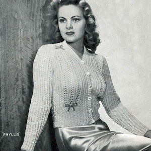 Puede incluir: Primer plano de una mujer con un cárdigan blanco de punto con cierre de botones y dos pequeños lazos en la parte delantera. El cárdigan está hecho con un delicado patrón de punto. La mujer lleva una falda y tiene el pelo peinado en suaves rizos.